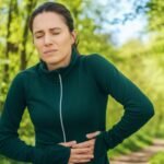 A woman in a green jacket clutches her stomach with a pained expression on a forest path.