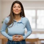 A smiling woman in a light blue long-sleeve polo shirt and jeans holds her hands over her stomach indoors.